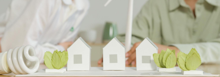 Three small white house models lined up on a table with green leafy plants on either side, and a coiled compact fluorescent bulb to the left, suggesting energy-efficient housing on a planning table with blurred people in the background.